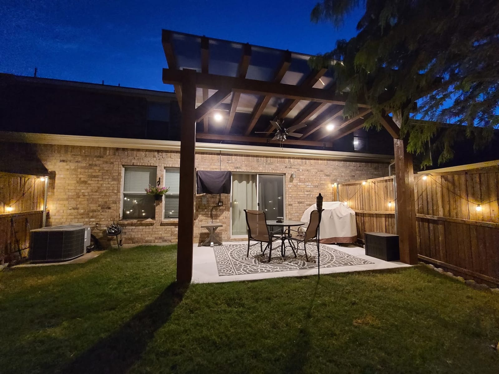 A backyard pergola with post and overhead string lighting glowing at dusk
